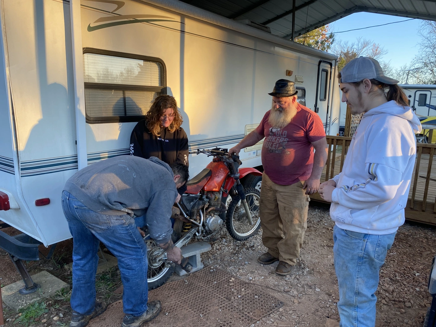 Four people working on a dirtbike in front of a camper