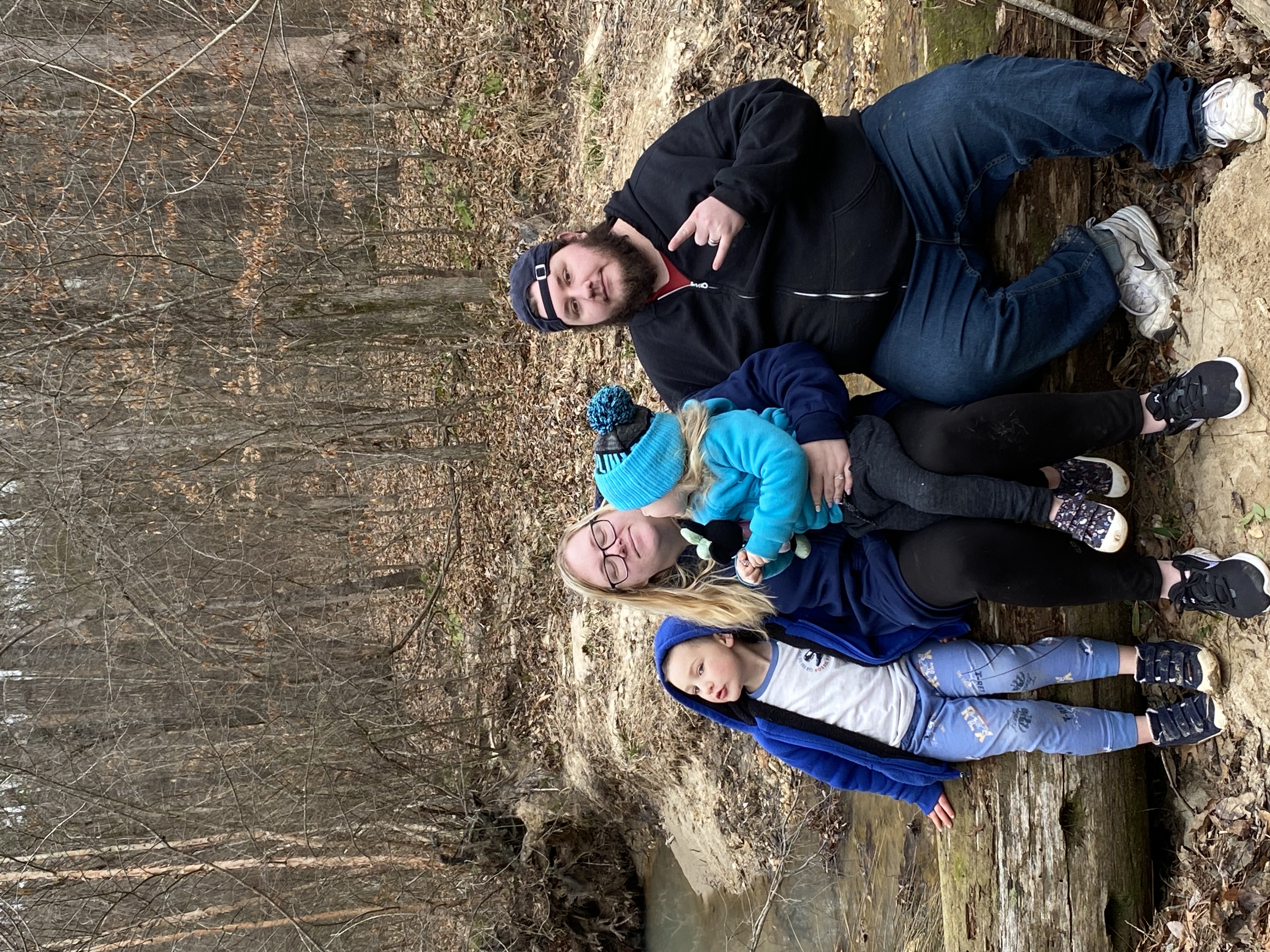 Two adults and one child sitting on a log, posing in front of a creek